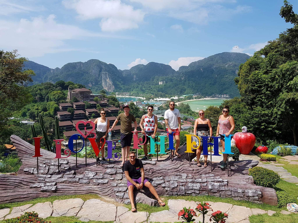 People posing with a large sign reading 'I Love Phi Phi' with hills in the background.