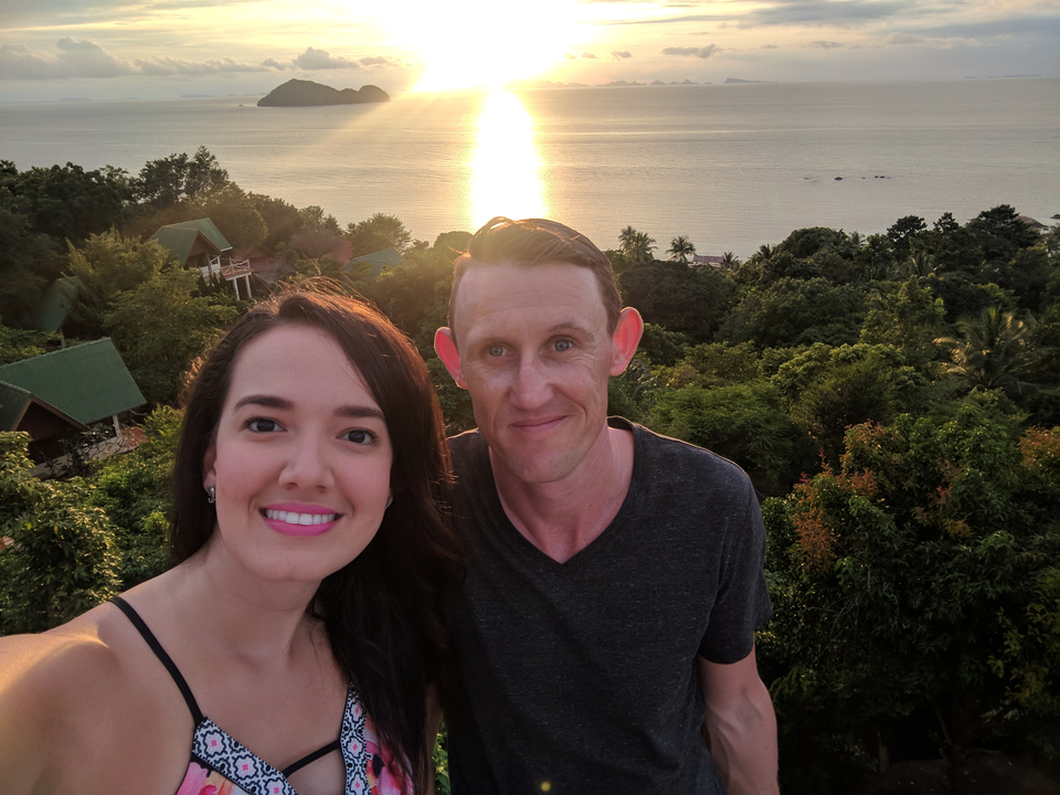 Two people taking a selfie with a sunset over a forested area.