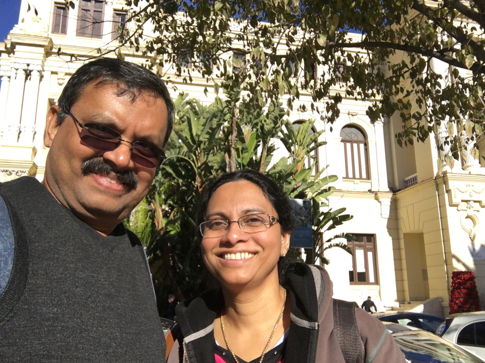 Deux personnes posant devant un bâtiment jaune avec un feuillage tropical.