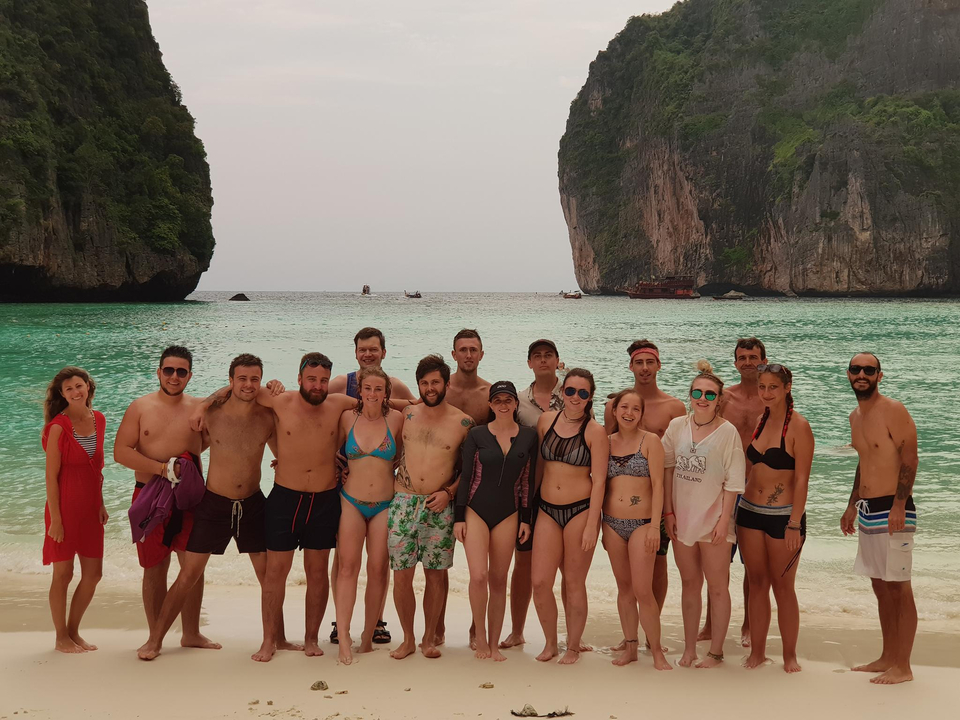 Large group posing on a beach with cliffs in the background.