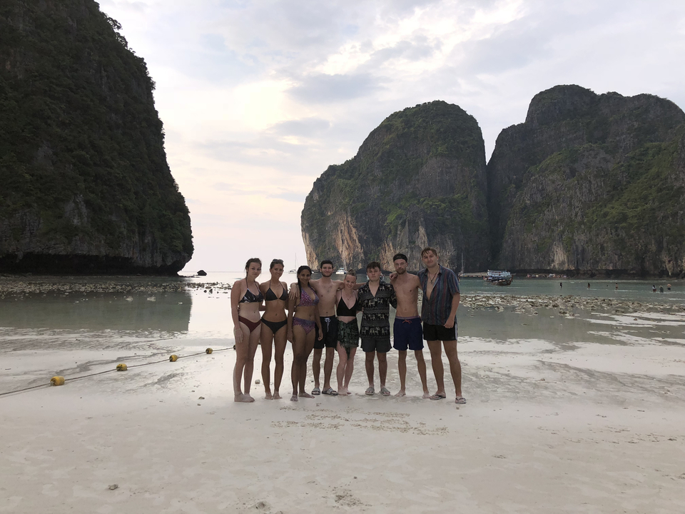Group of people standing on a beach with large rock formations in the background.