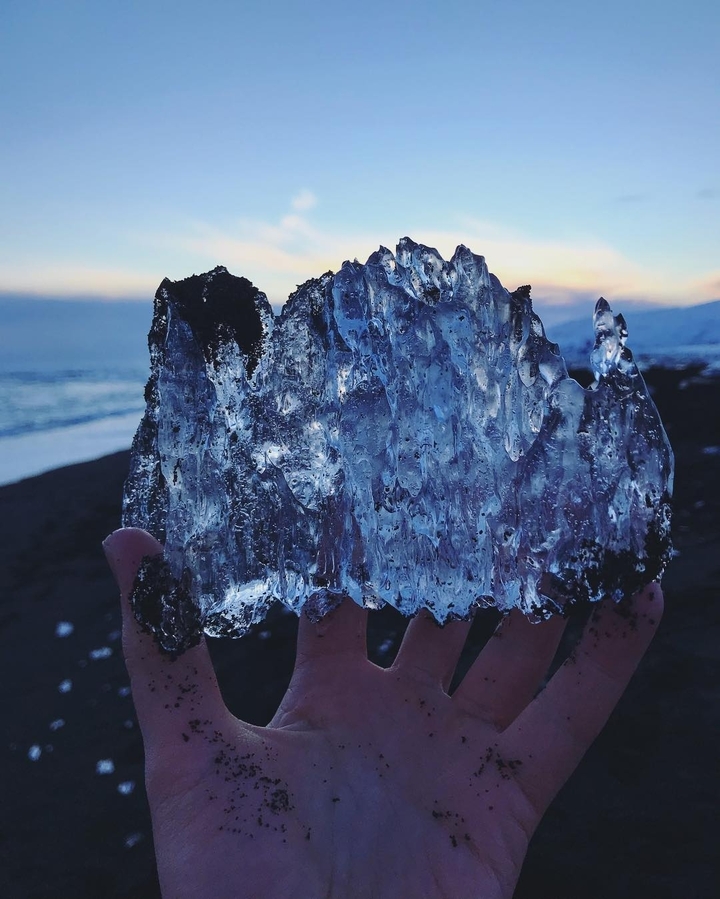 Person holding a large piece of ice on a black sand beach.