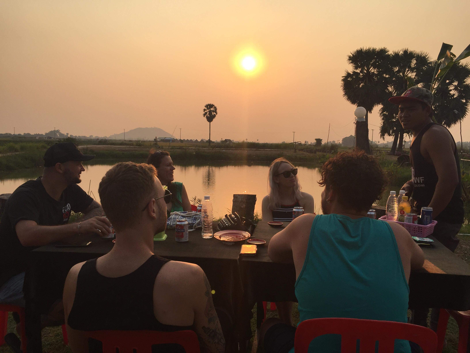 Group of people sitting by a lake while the sun sets.