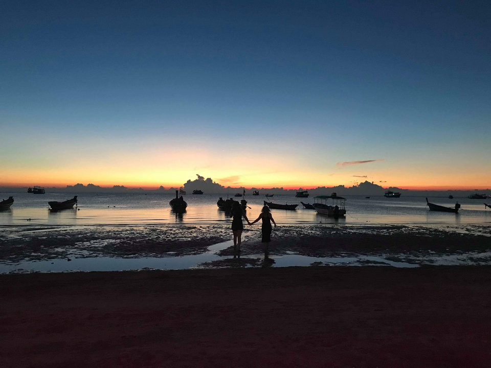 Silhouette of two people at a beach during sunset with boats in the background.