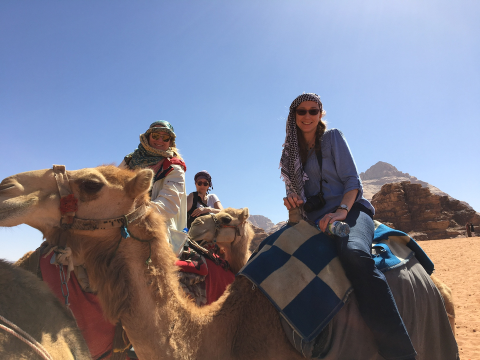 Three people riding camels in a desert with rocky formations in the background.