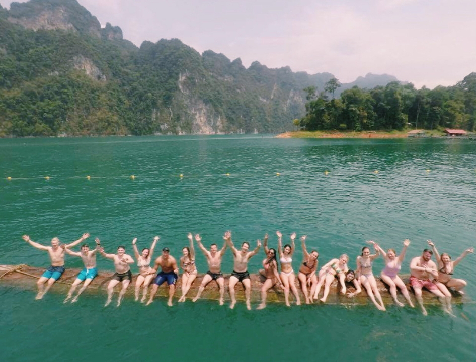 Group of people sitting on a log in a lake with mountains in the background.