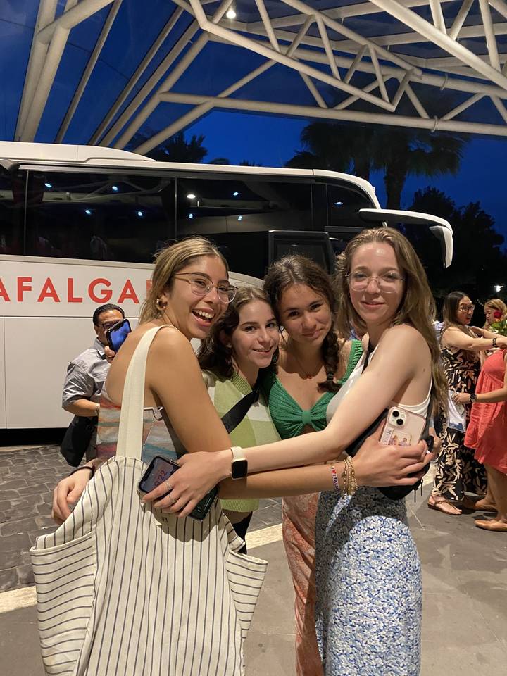 Group of friends hugging outside a building, possibly a travel bus nearby.