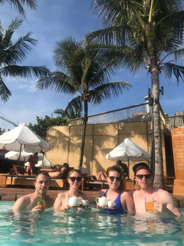 People relaxing by poolside with palm trees and umbrellas.