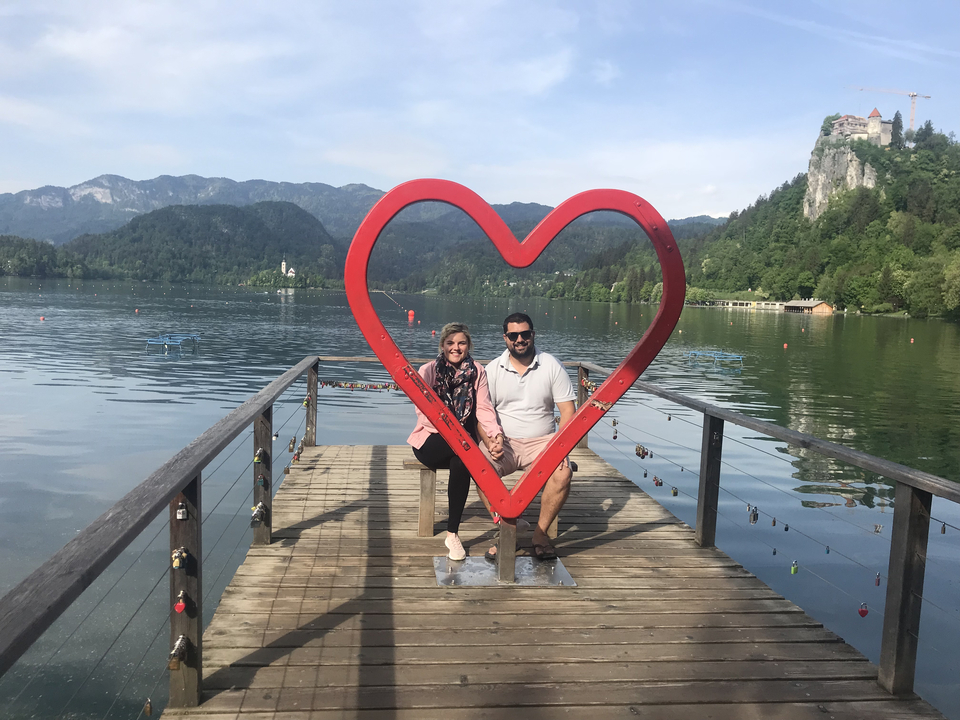 Couple posing with a large red heart sculpture on a dock over a lake.