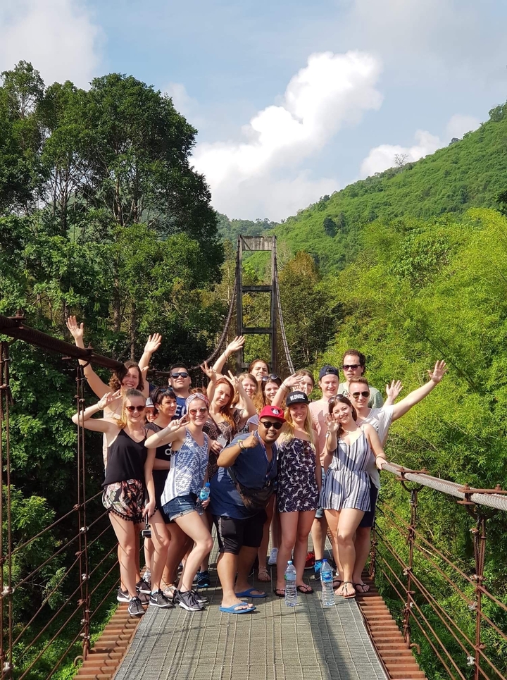 Group posing in front of a suspension bridge and lush green trees.