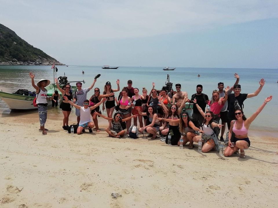 Group posing on a sandy beach with boats in the background.