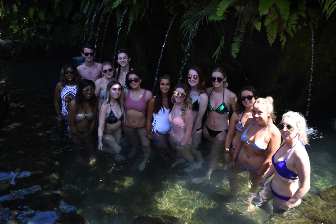 Group of young people standing in shallow water under a waterfall.