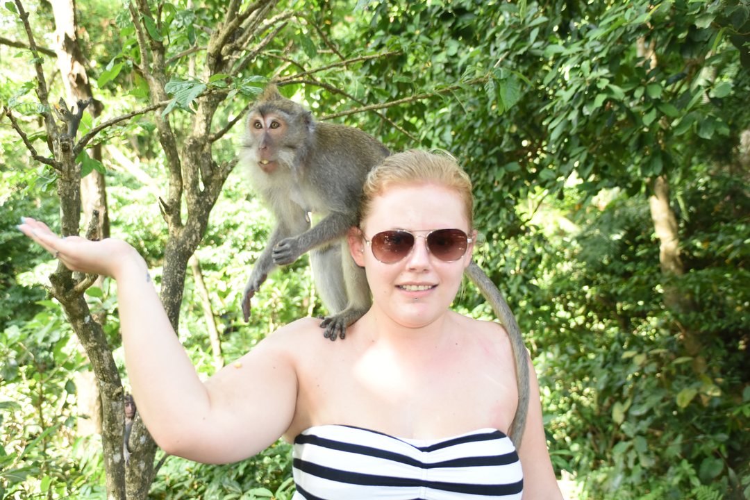 A person with sunglasses posing with a monkey on their shoulder in a jungle setting.
