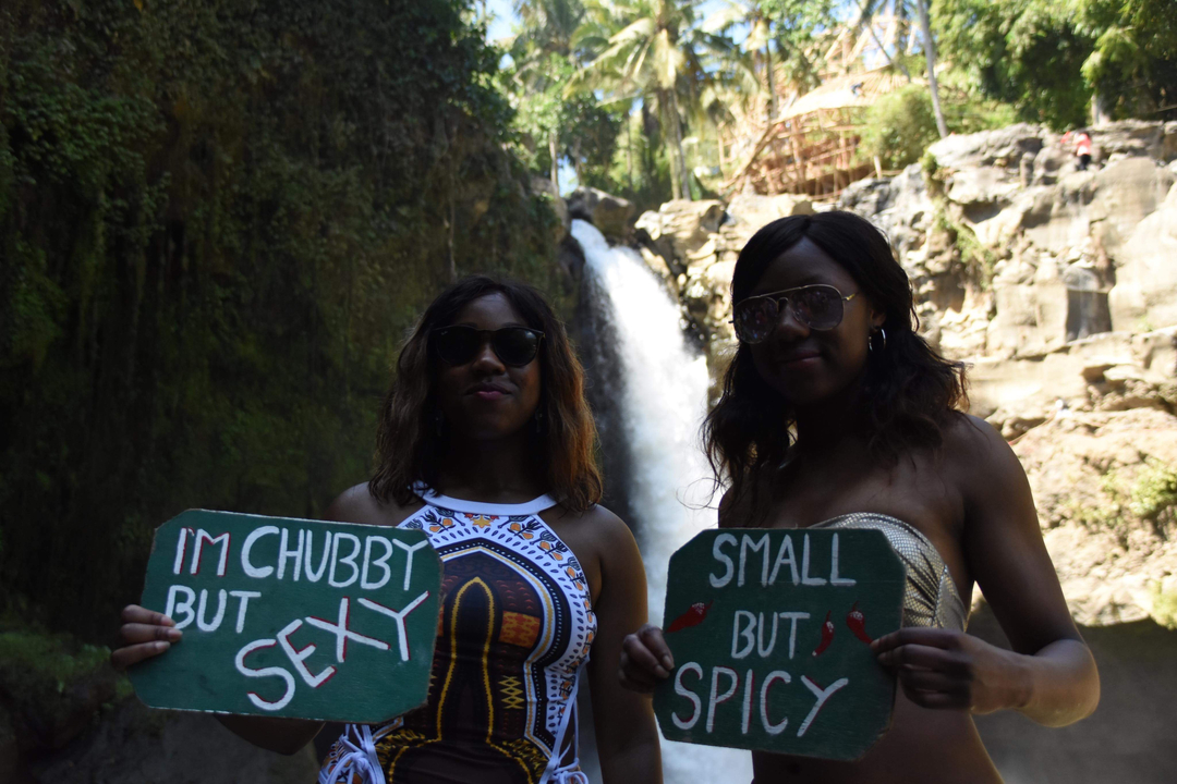 Two women posing with signs in front of a waterfall.