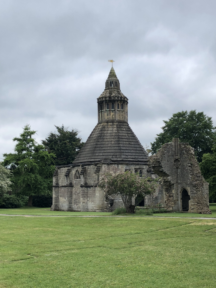 A stone chapel with a conical roof among ruins and greenery.