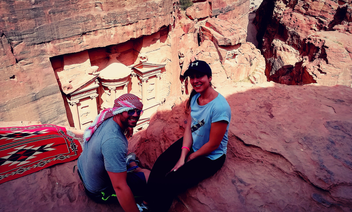 Two people sitting on a rock ledge overlooking the Petra Treasury.