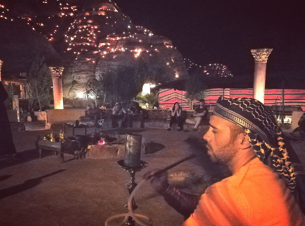 Man smoking a hookah outside with fire-lit mountains in the background.