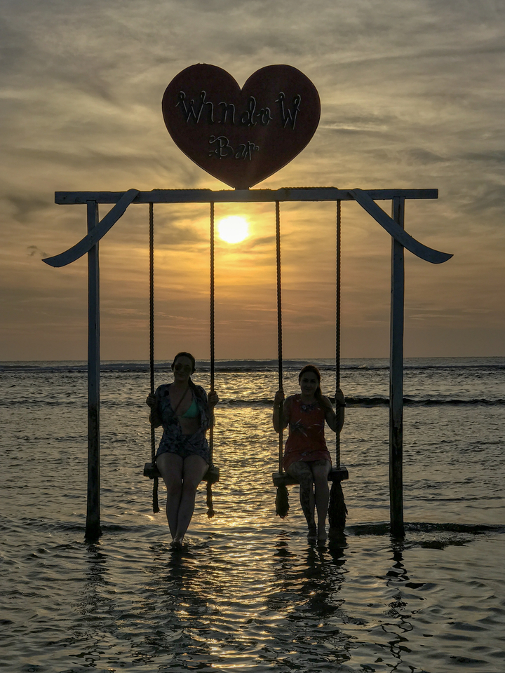 Two people on a swing in front of a sea with sunset in the background.