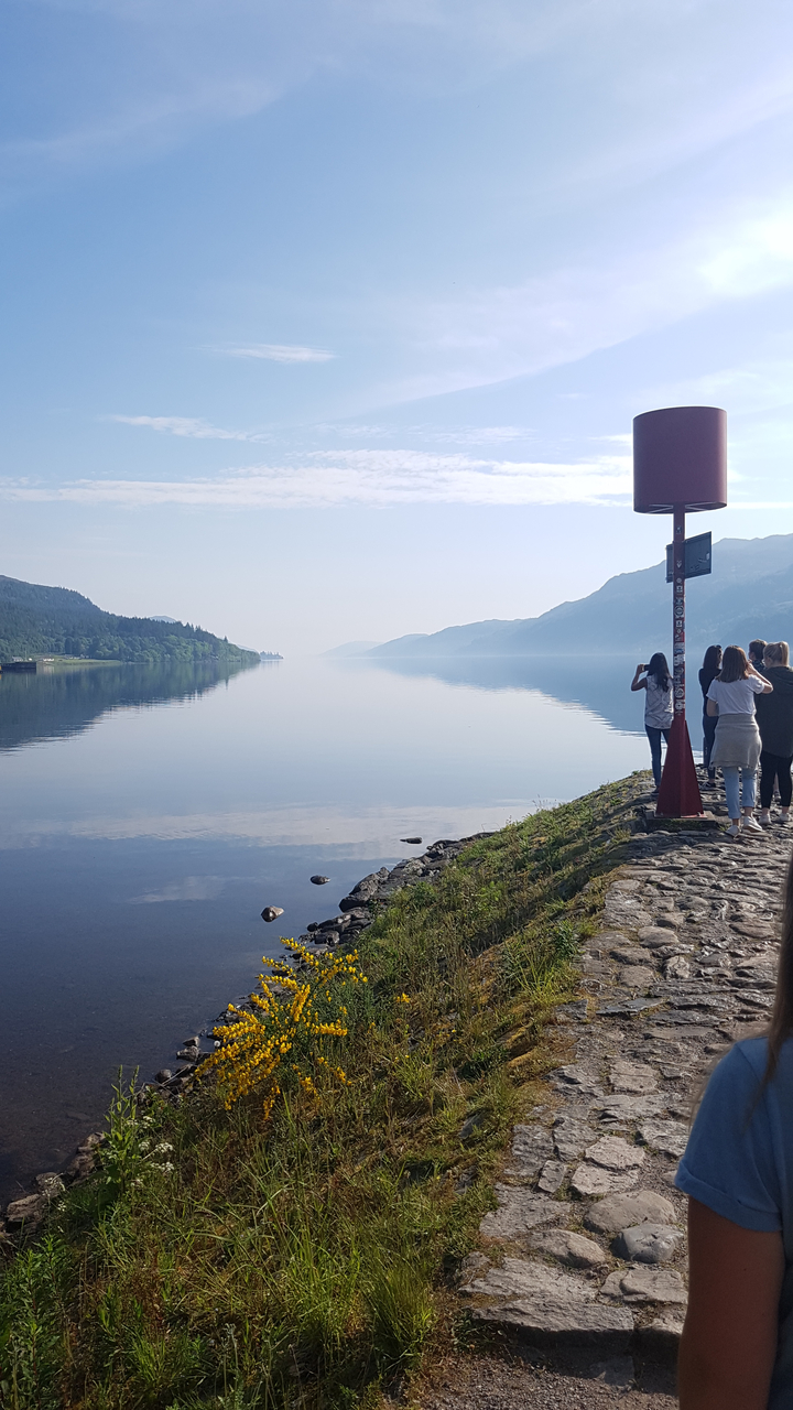 People taking photos by a calm lake with mountains in the distance.