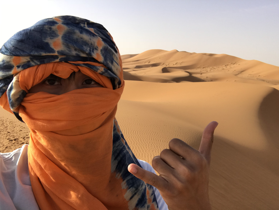 Person in traditional attire in the desert with sand dunes.