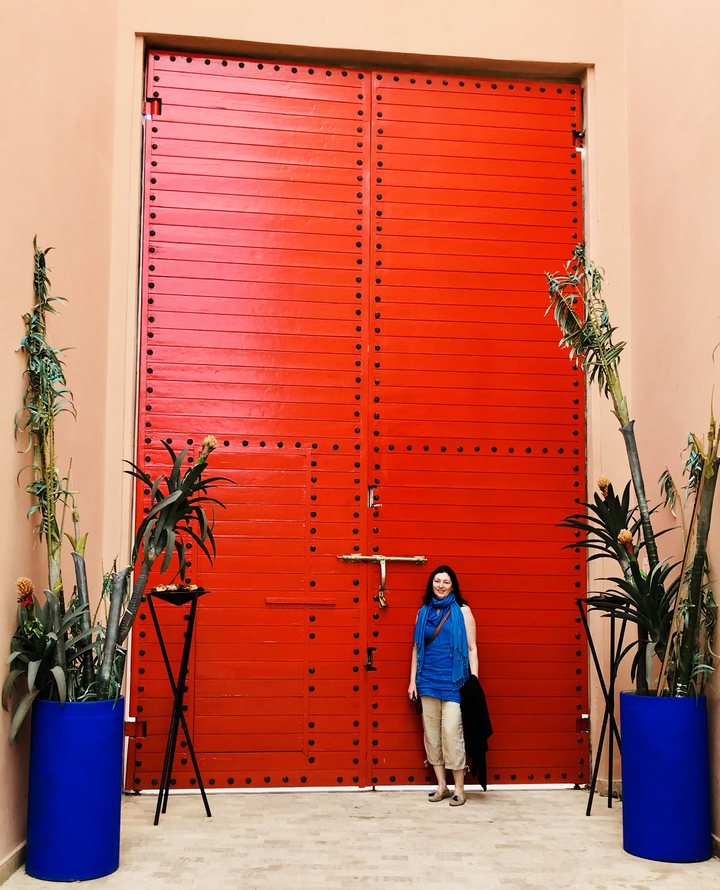 A woman in front of a massive red door surrounded by plants.
