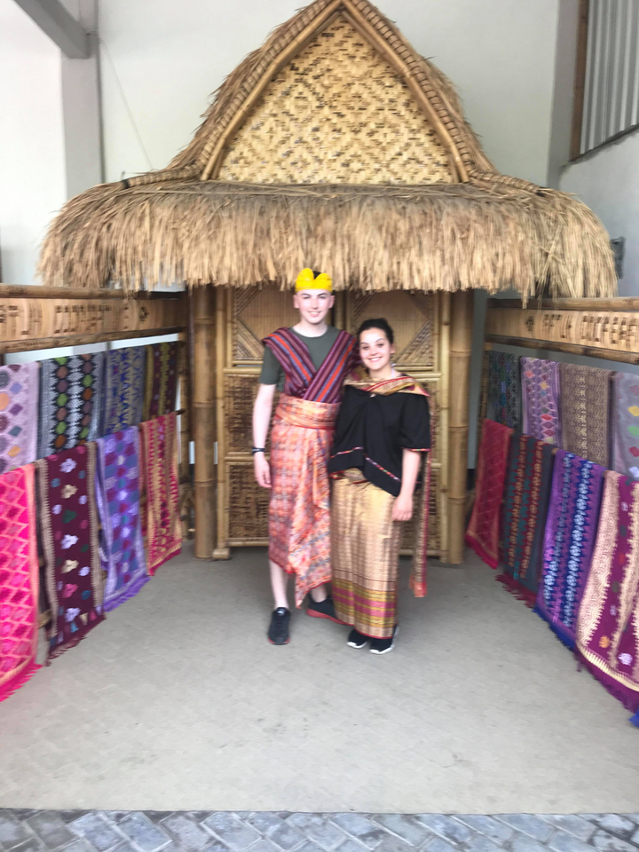 Couple dressed in traditional attire standing amidst colorful textiles.