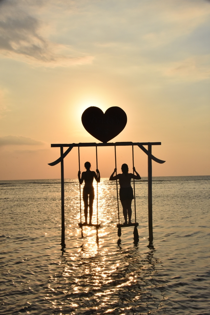 Two people sitting on swings in front of a silhouette heart shape at sunset.