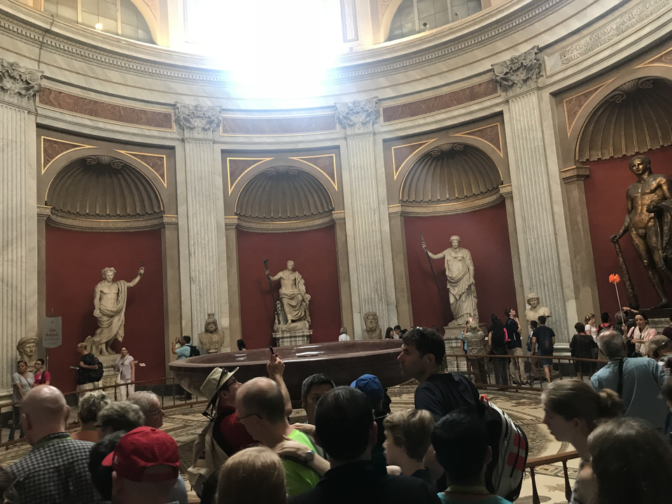 Interior of a building with classical statues and a red wall, crowded with visitors.