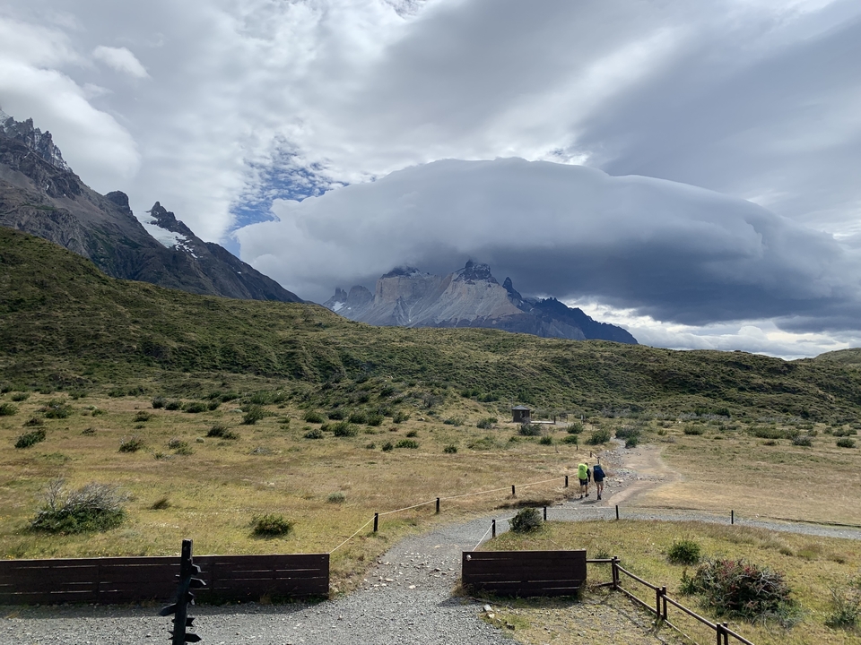 Two people hiking a trail with dramatic cloud-covered mountains.