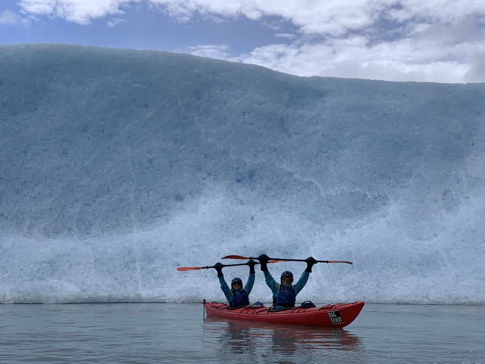 Two kayakers salute with paddles by a massive glacier wall.