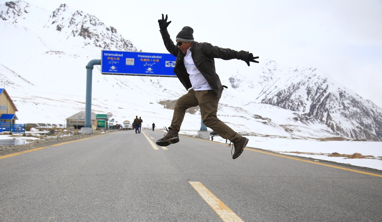 Person jumping on a snowy road with mountains in the background.