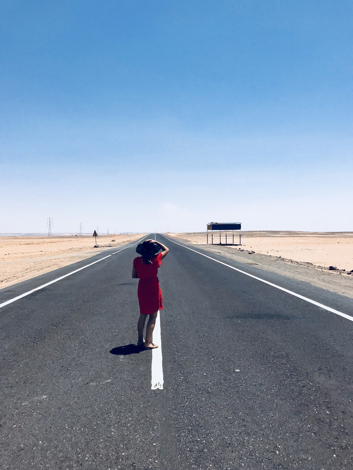 Woman in a red dress standing on an empty desert highway stretching into the horizon.
