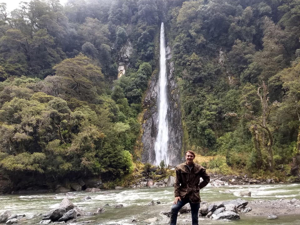 Man posing in front of a tall waterfall.