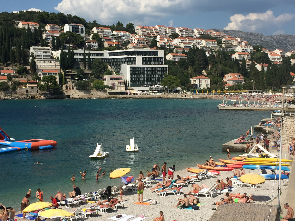 Beachfront area with people sunbathing and swimming, with a distant view of buildings along the shoreline.