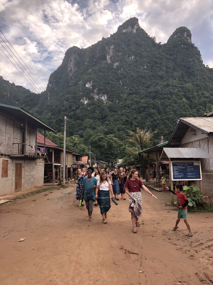 Group walking through a village with lush vegetation and hills in the background.
