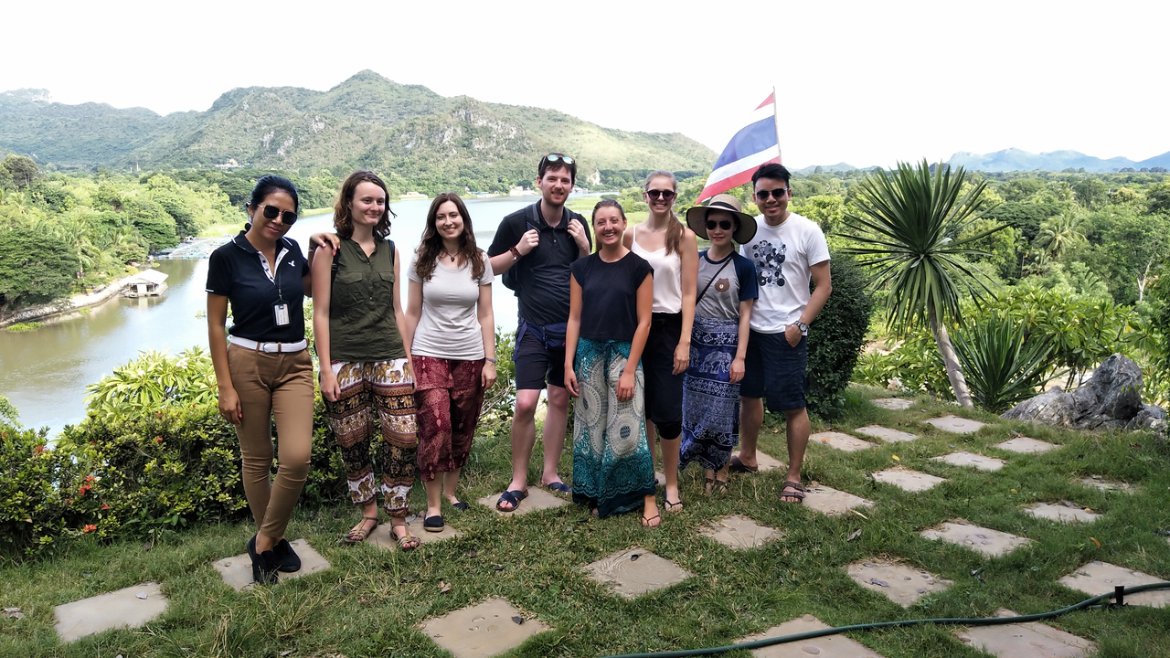 Group posing with a Thai flag and scenic landscape.