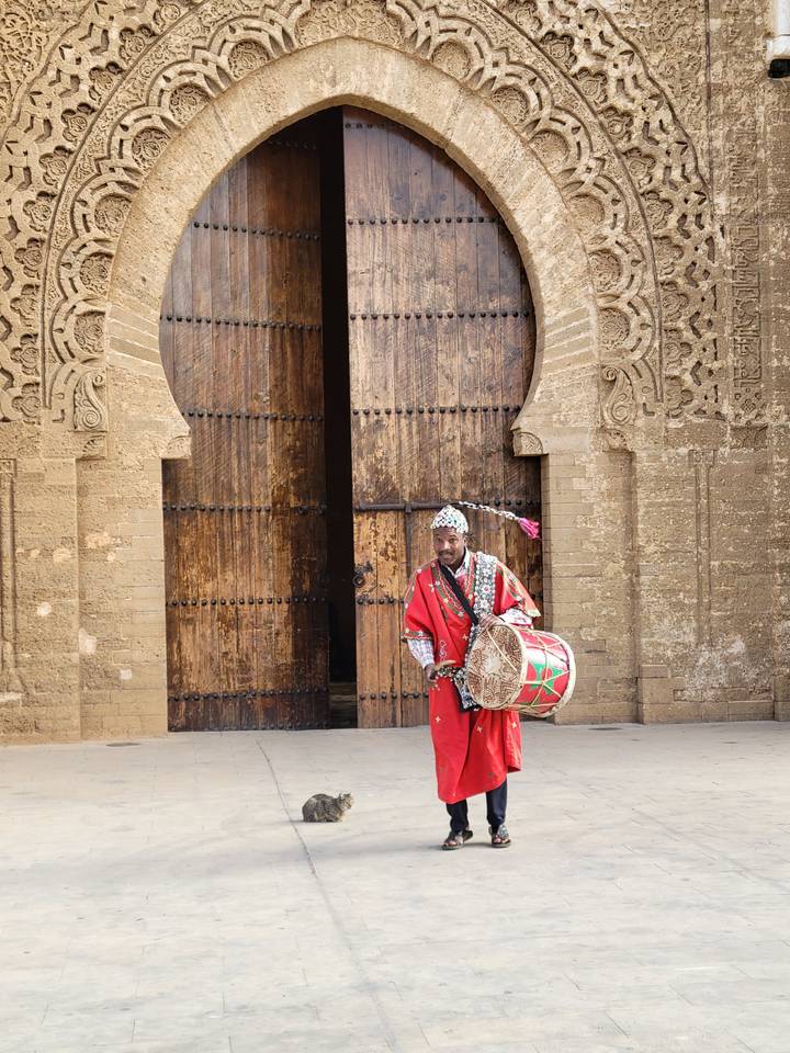 Performer in traditional outfit playing a drum outside a large arched doorway.