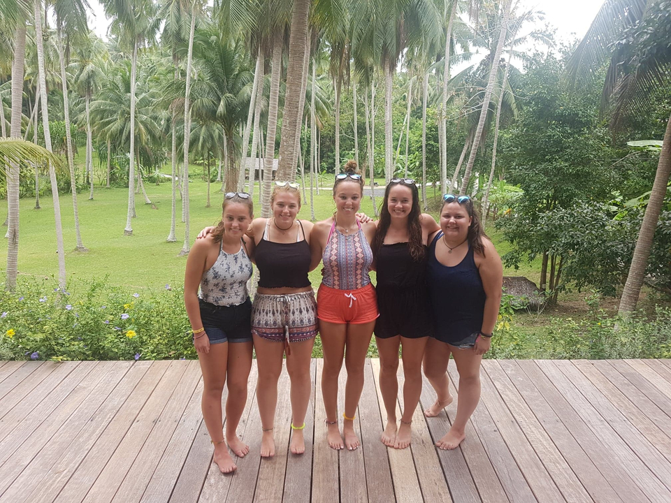 Five young women standing on a wooden deck surrounded by palm trees.