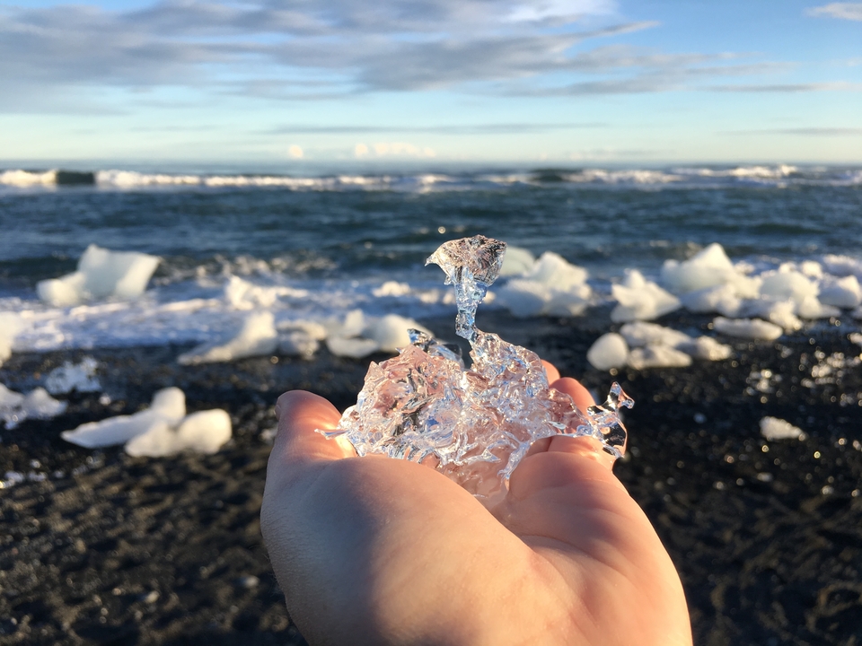 Hand holding a piece of ice on a black sand beach with ocean.