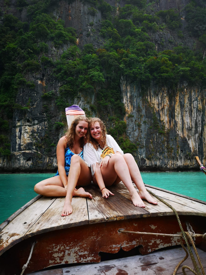 Two women sitting on a boat in front of rocky cliffs and turquoise water.