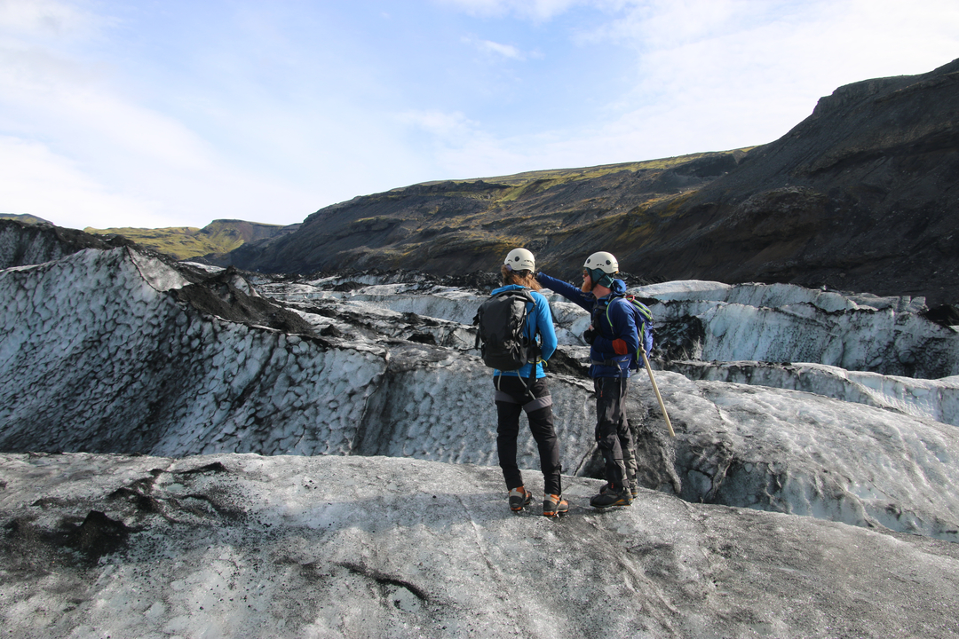 Two people wearing outdoor gear standing on a glacier pointing at the landscape.