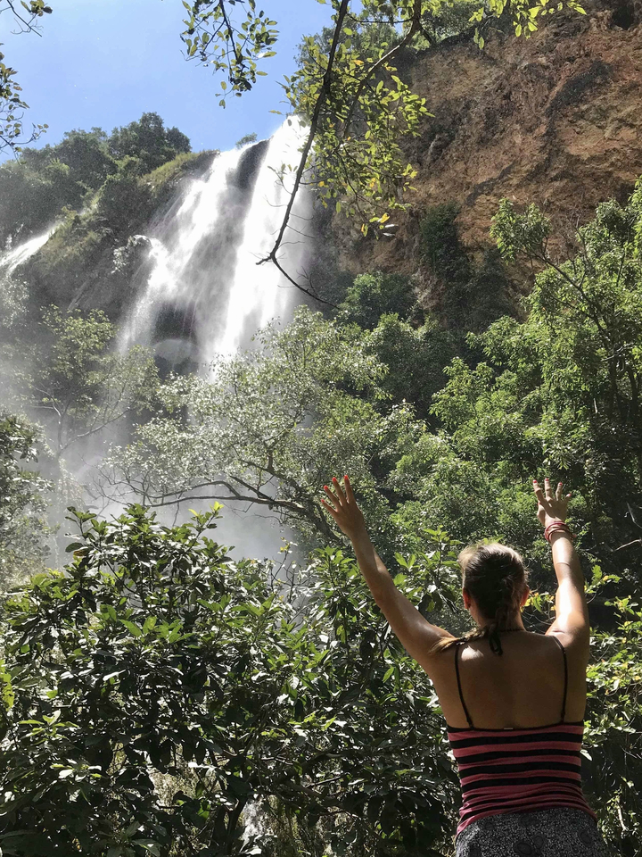 A woman with arms raised, looking at a waterfall through trees.
