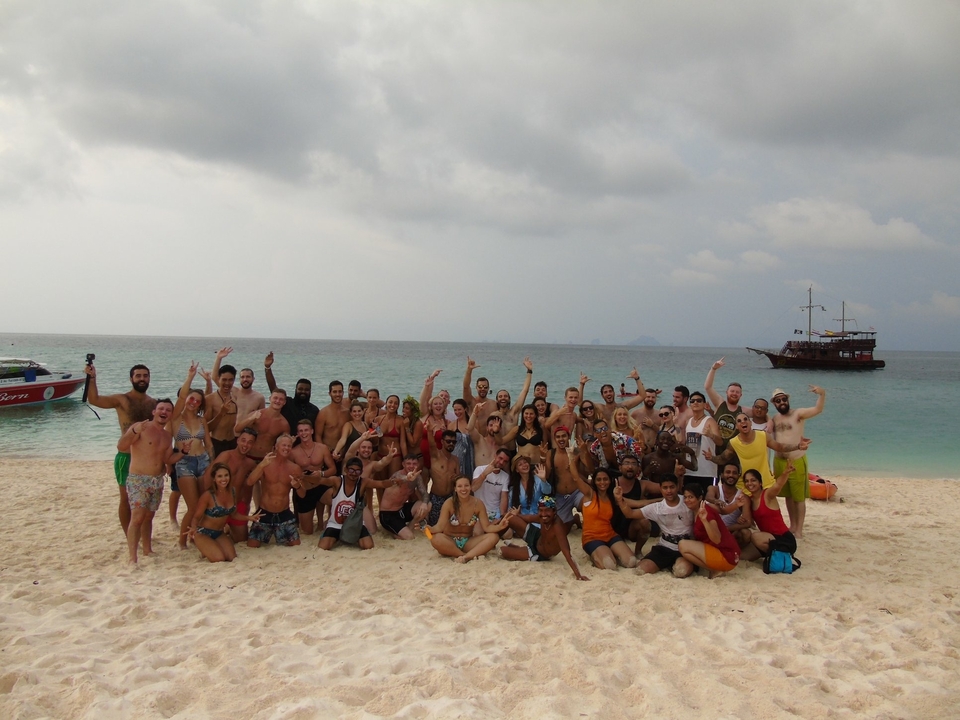 A large group of people on a tropical beach with a ship visible.