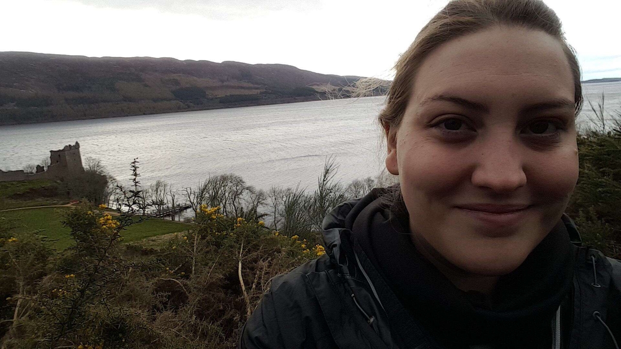 Person taking a selfie with a lake and ruins in the background.