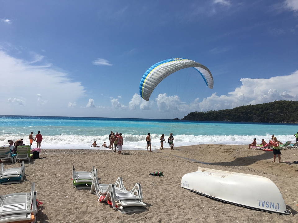 Paragliding over a vibrant blue sea with people relaxing on a sandy beach.