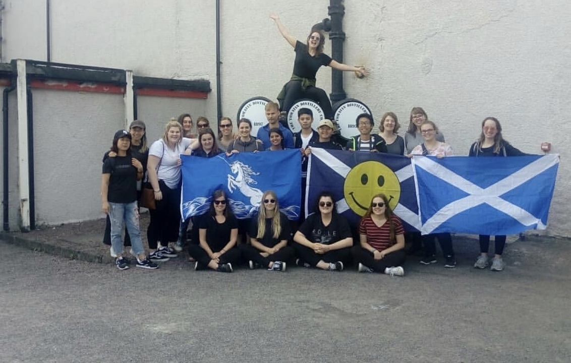 A large group photo in front of a distillery with flags.