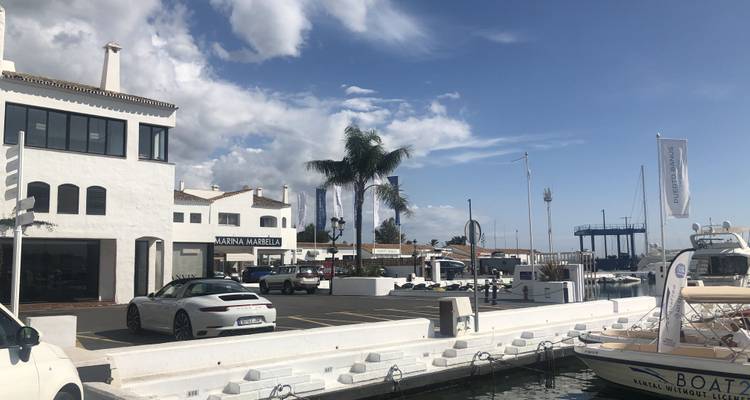 Marina avec voitures de luxe et bateaux, ciel dégagé en arrière-plan.