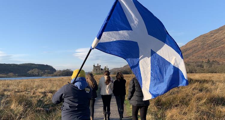 Personas con una gran bandera escocesa caminando hacia las ruinas de un castillo.