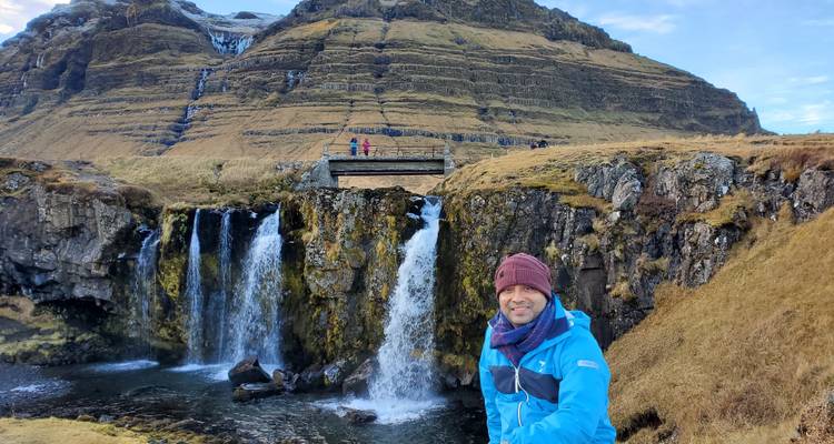 Smiling person in front of scenic waterfalls and mountains.