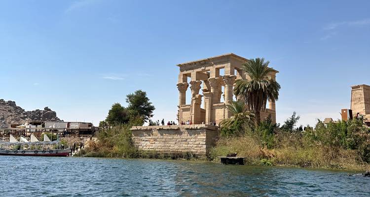 Ruinas de un templo antiguo en la orilla de un río con palmeras y turistas cerca.